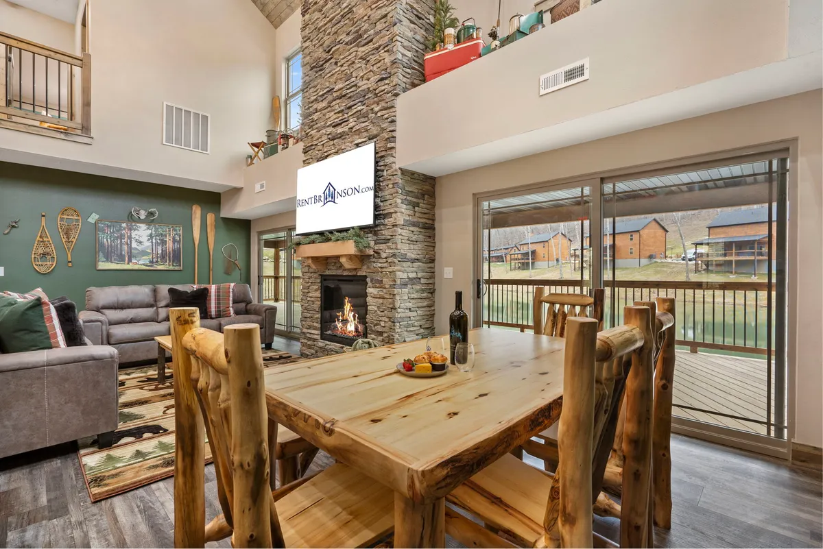 A spacious open-concept interior featuring a stone fireplace as the focal point, a large wooden dining table, comfortable seating area, expansive windows filling the space with light, and decorative wall art. The scene conveys warmth and togetherness, suggesting a perfect spot for gathering, storytelling, and sharing meals.