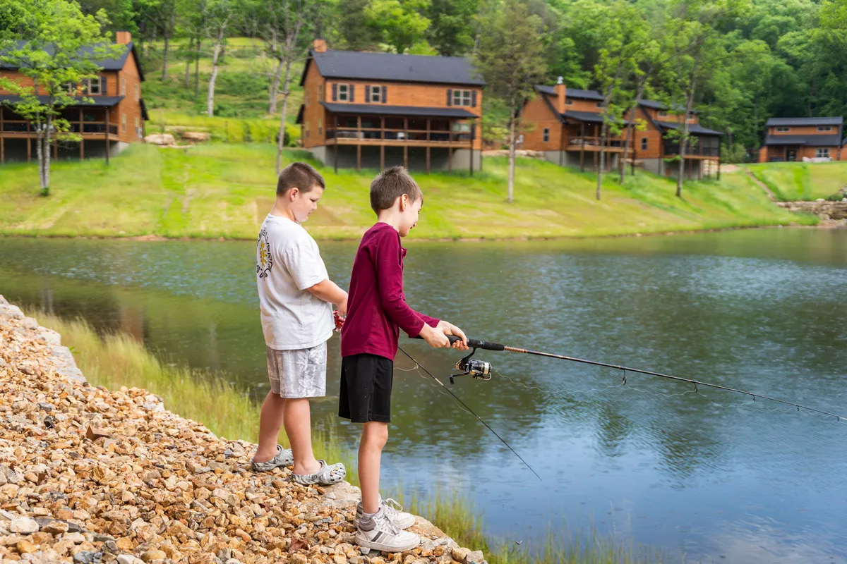 Two boys fish at a tranquil lake, with wooden cabins and serene nature in the background. The scene is playful yet peaceful, suggesting family bonding and the unique opportunity to relax by the water's edge within the resort community.
