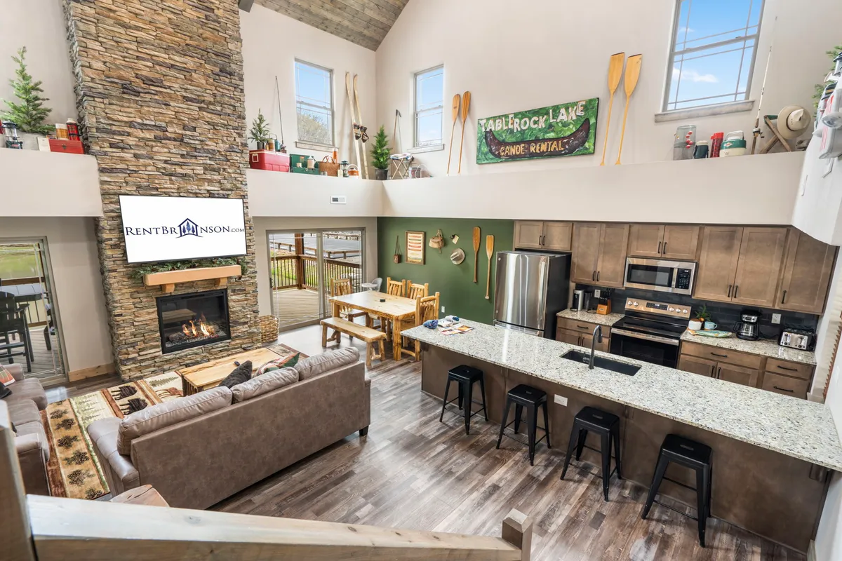 The image showcases the inviting interior of a cabin living space, featuring a grand stone fireplace as the centerpiece, surrounded by comfortable seating. Expansive windows let in natural light and offer tranquil water views, while a spacious kitchen island and dining table set the stage for family meals and gatherings. The scene radiates warmth, blending rustic charm with modern comforts, and hints at memorable moments—whether relaxing by the fire, sharing holiday dinners, or enjoying laughter-filled games.
