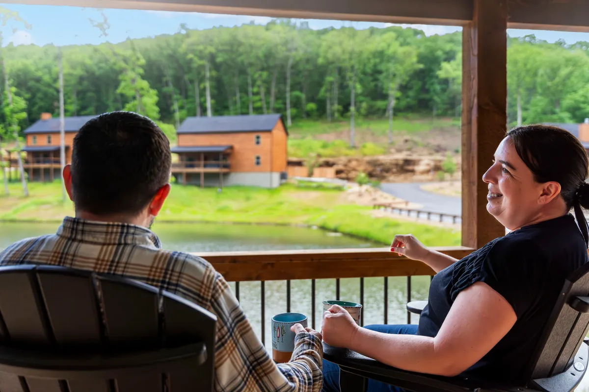 Two people sit comfortably on a porch with mugs in hand, looking out across a serene lake with cabins in the background. The mood is cheerful and intimate, perfect for sharing a morning coffee or reflecting at sunset.