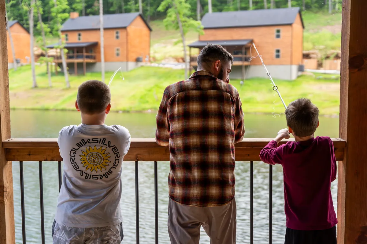 This image captures a family-friendly scene with fishing rods by a picturesque body of water, surrounded by wooden cabins and lush green trees. The atmosphere is joyful and serene, emphasizing outdoor activities like fishing and relaxing by the water.
