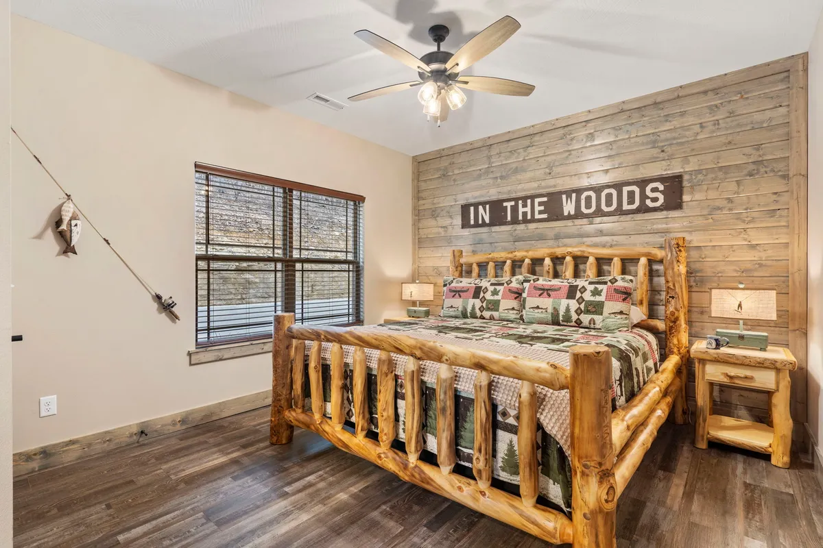 A cozy bedroom featuring a log bed, wood-paneled walls, a decorative sign, and a window with blinds that lets in natural light. The bedding appears plush and inviting, with thoughtful rustic accents creating a restful atmosphere.