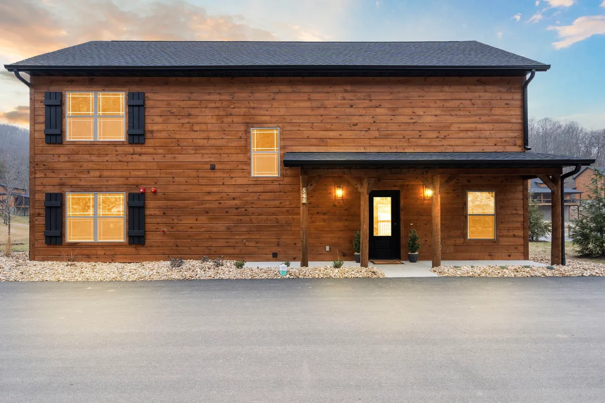 This image features a cozy wooden cabin with a welcoming front porch, illuminated windows, and lush shrubbery, all set against a tranquil natural backdrop. The porch invites relaxation and social moments, while the warm lighting hints at comfortable gatherings inside.