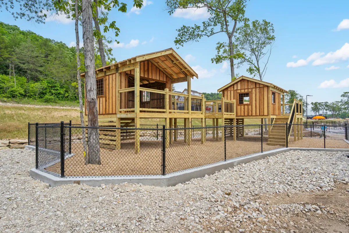 The image showcases vibrant playhouses set among trees and a gravel area, with children at play and families gathered close by. The setting is energetic and filled with the joy of kids exploring and parents enjoying time outdoors.