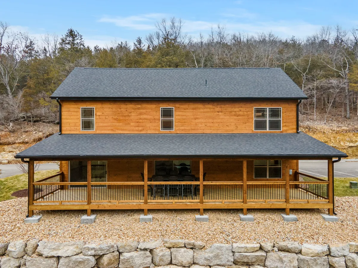 A welcoming exterior view of Pop’s Fishing Cabin with warm wooden siding, a spacious porch, and large windows that glow in the light. The stone foundation and inviting porch hint at evenings spent enjoying sunset views or mornings sipping coffee outdoors. The surrounding nature and ample porch space suggest opportunities for gathering, relaxing, and outdoor dining.
